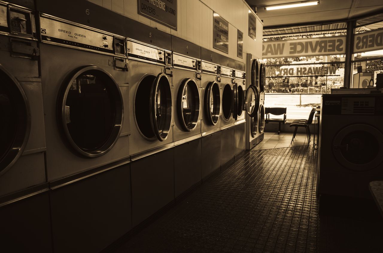 about-img-01 Sepia-toned image of a laundromat interior showcasing a row of classic washing machines.